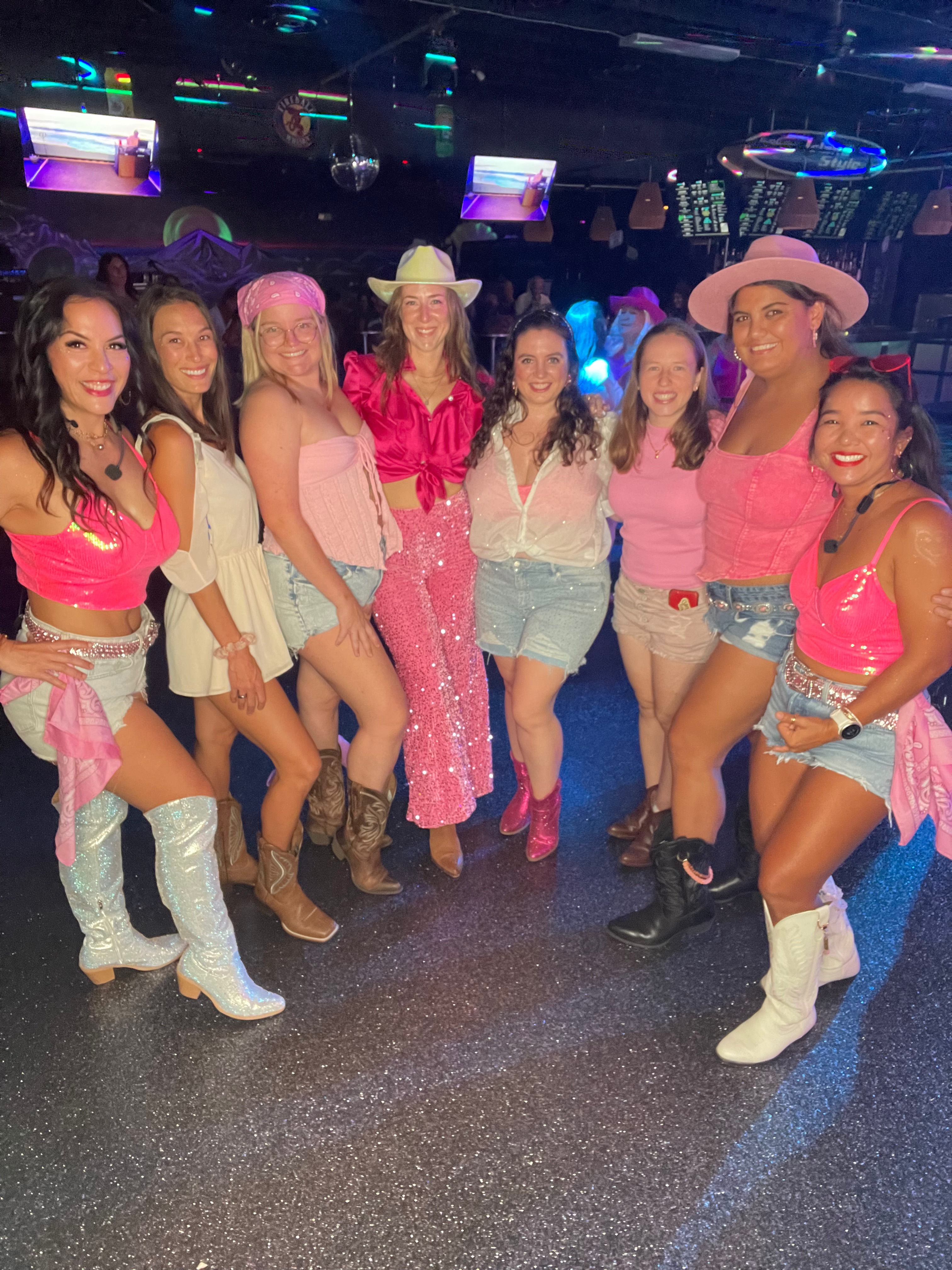A group of Bootscootin Maui dancers in pink and denim, laughing together on the dance floor