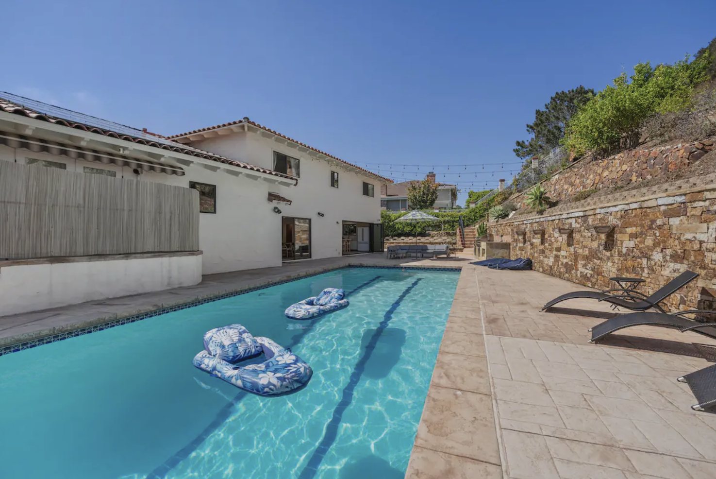 The swimming pool seen along its full length, with two floats in the water, a stone retaining wall and lounge chairs
