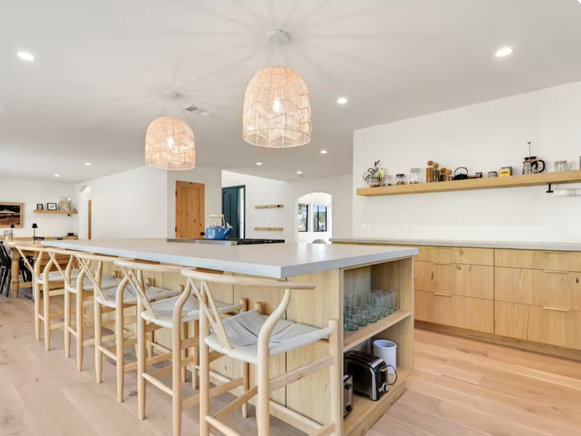 A kitchen with a long wood island, woven pendant lights and wishbone stools, opening to a dining area