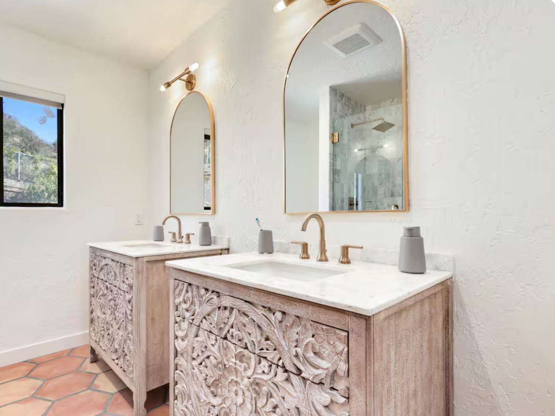 A bathroom with two carved wood vanities, arched gold mirrors and a window