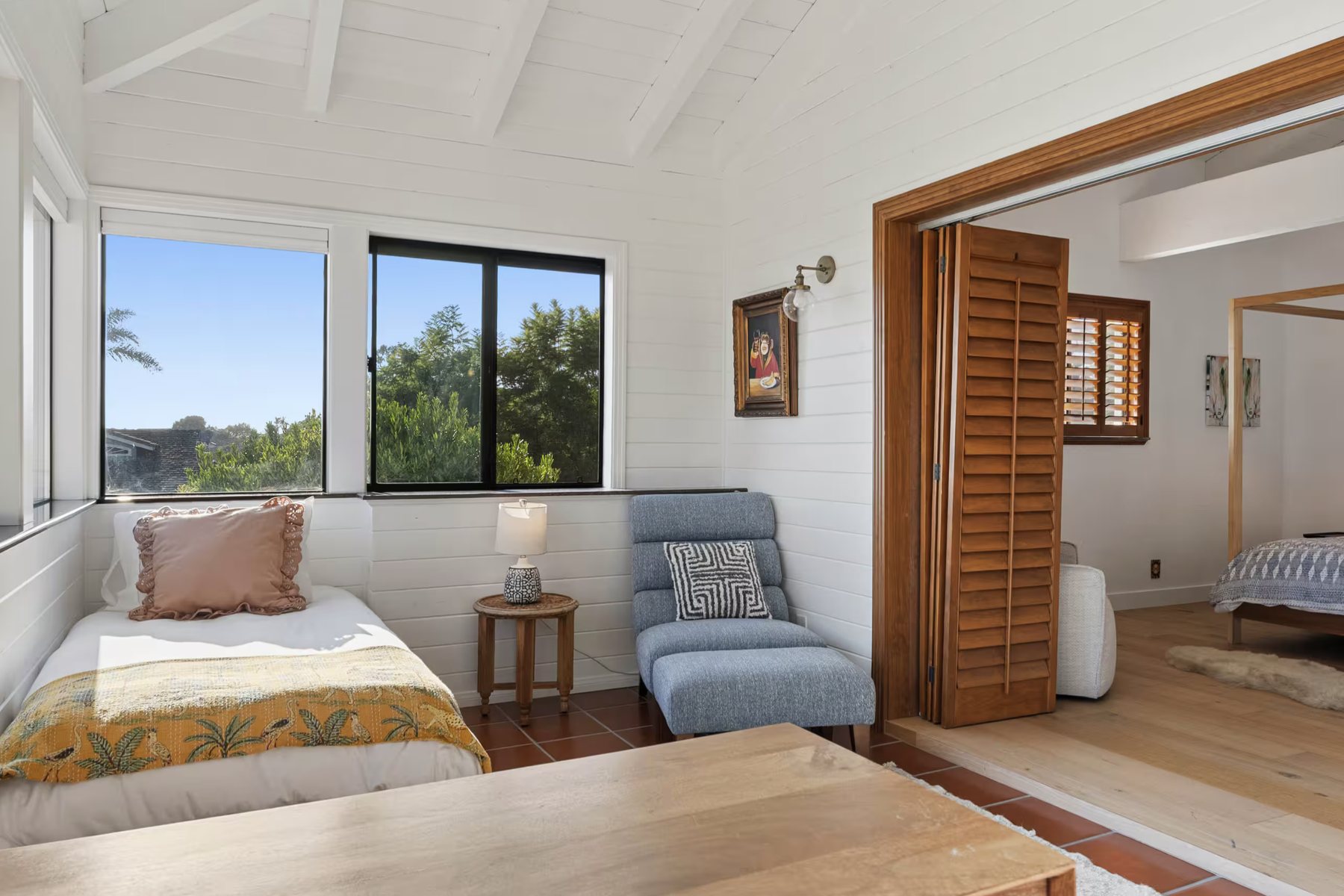 A sunny room with a daybed, a blue lounge chair and ottoman, and windows with garden views