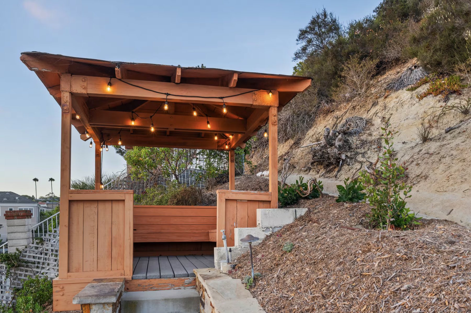 A wooden meditation gazebo with string lights, a built-in bench and a stone step, set against a landscaped hillside